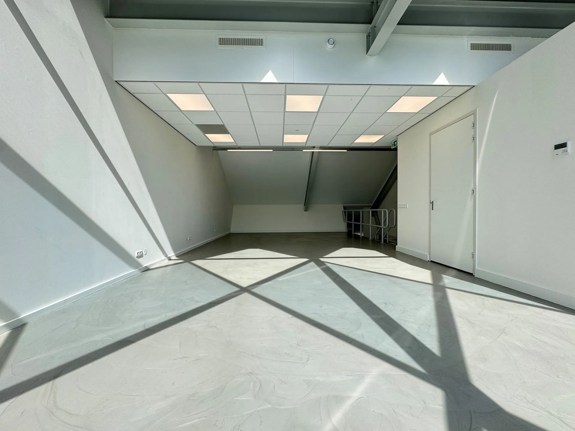 Lichte, lege ruimte in Mart Stamhof met betonnen vloer, systeemplafond en opvallende lichtinval door schaduwen.